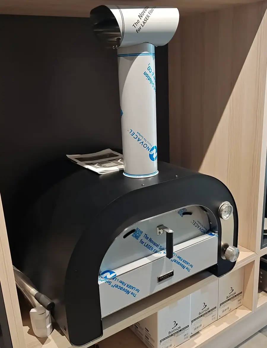 Brabura wood-fired pizza oven in matt black with stainless chimney, shown on a showroom shelf alongside Borniak smoker packaging