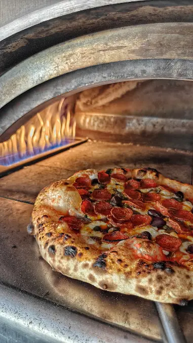 Wood-fired pizza with pepperoni, olives and melted cheese cooking in a pizza oven at Joe's Street Pizza