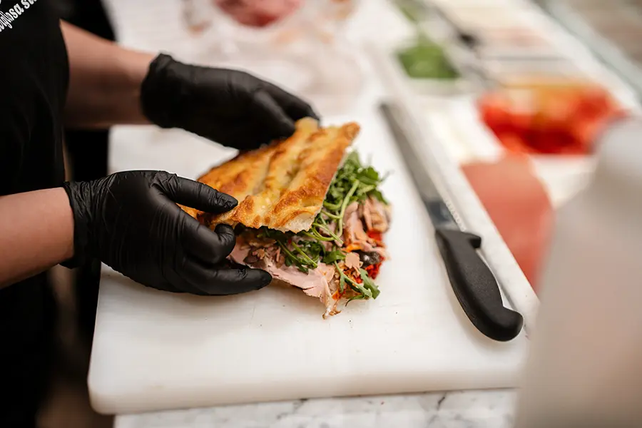 All'Antico Vinaio team member in black gloves holding a completed schiacciata sandwich wrapped in branded paper