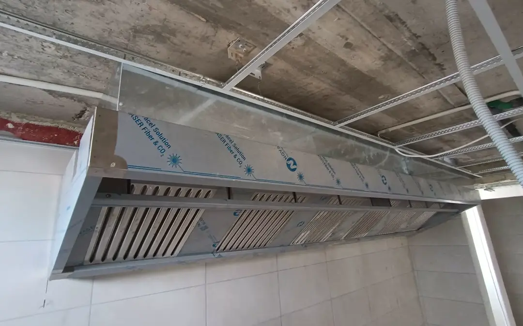 Wide extraction canopy spanning the full cooking line in a commercial kitchen under construction, viewed from below