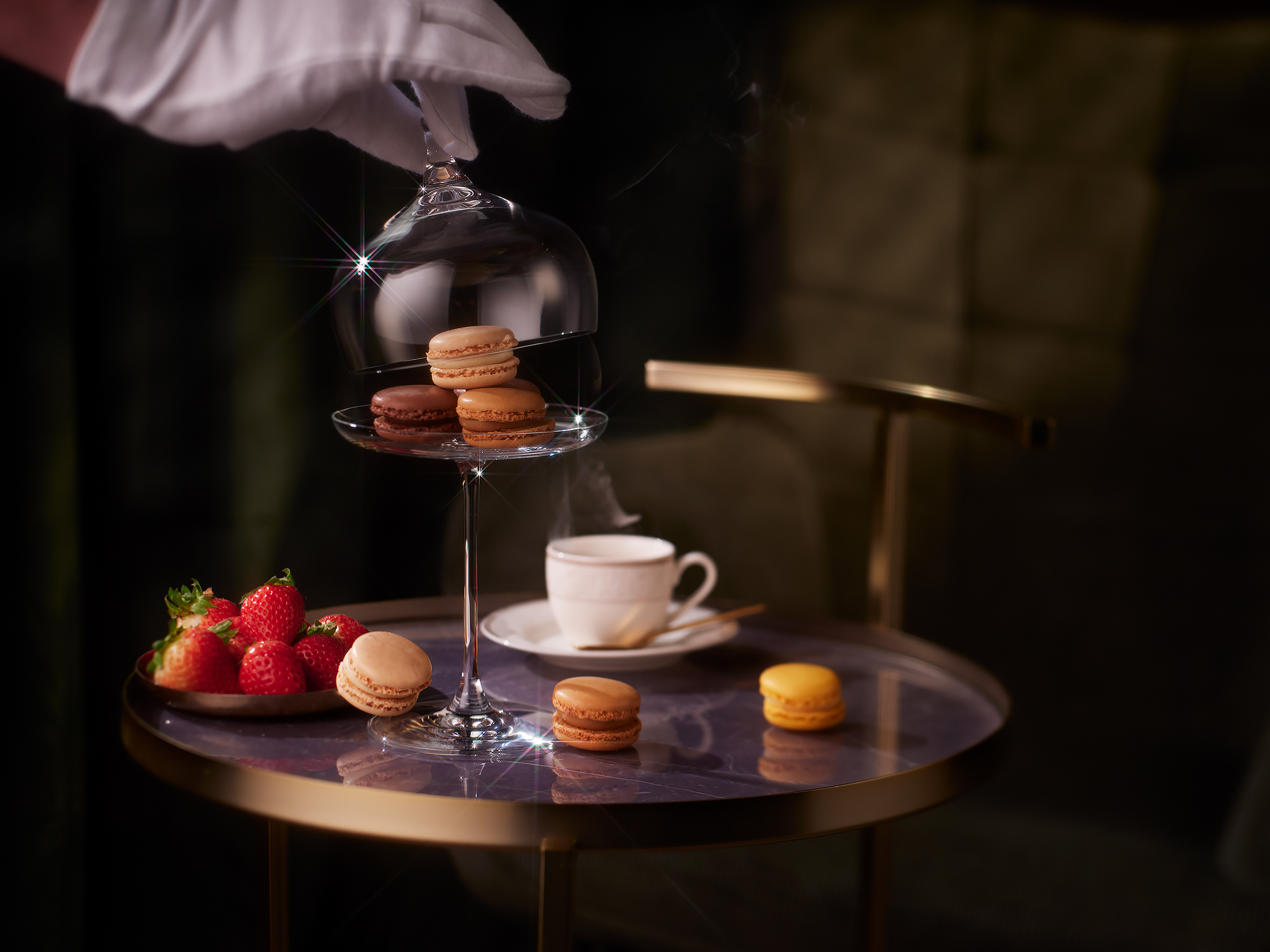 Rona crystal cloche lifted by a white-gloved hand to reveal macarons on a stemmed cake stand, with a steaming espresso cup on a marble-top side table