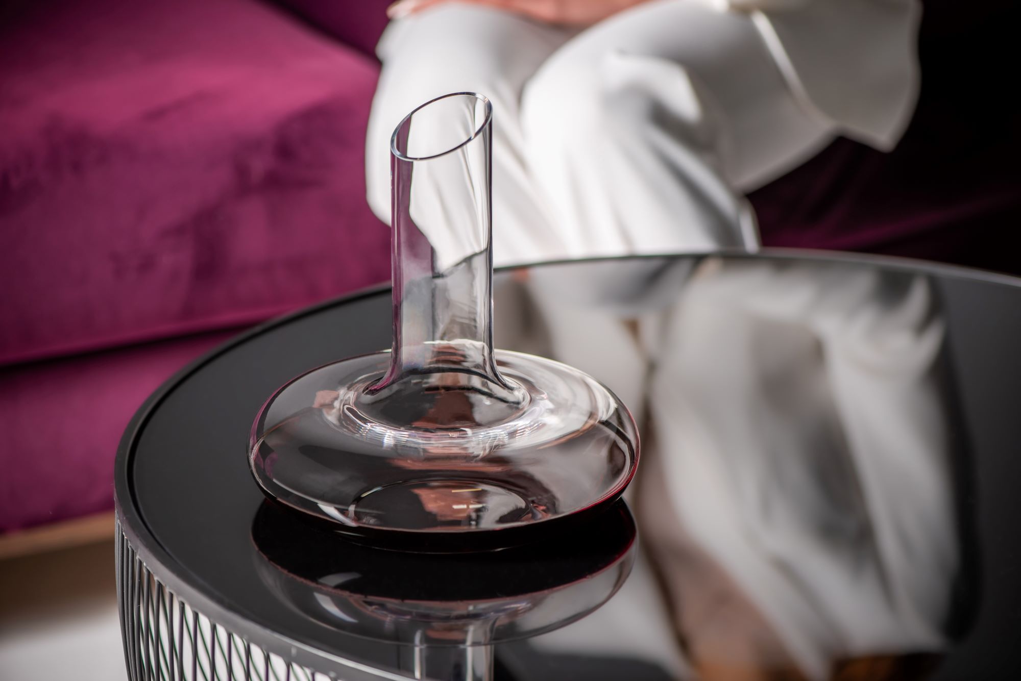 A Rona handmade lead-free crystal decanter with a wide flat base and straight neck resting on a dark side table, with a white-gloved server's hand visible