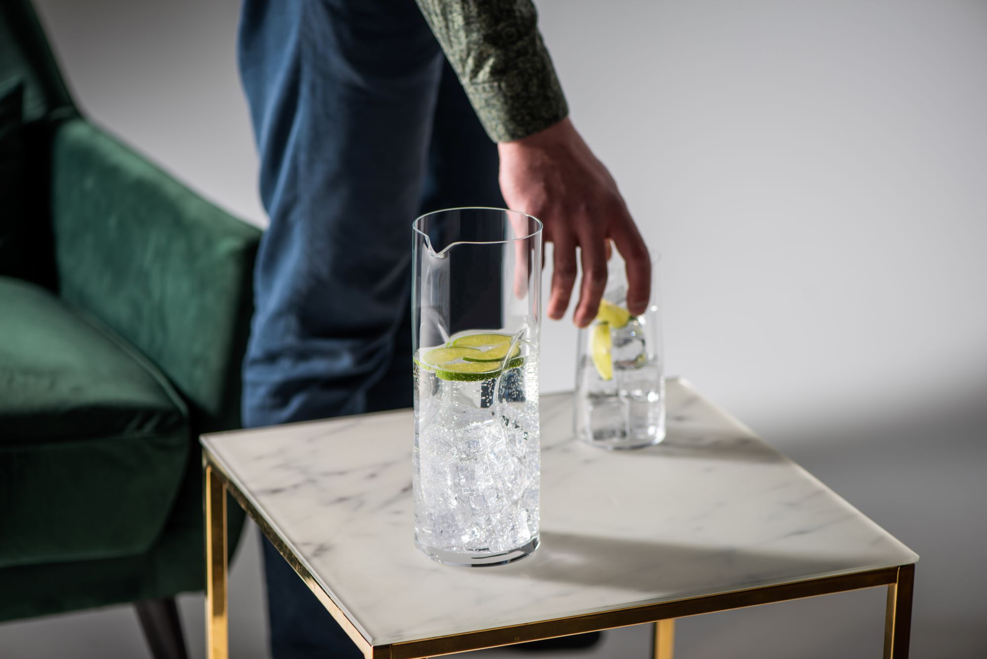 A Rona glass pitcher filled with iced water and lime slices on a marble-top brass side table, with a matching tumbler being lifted by a hand in the foreground