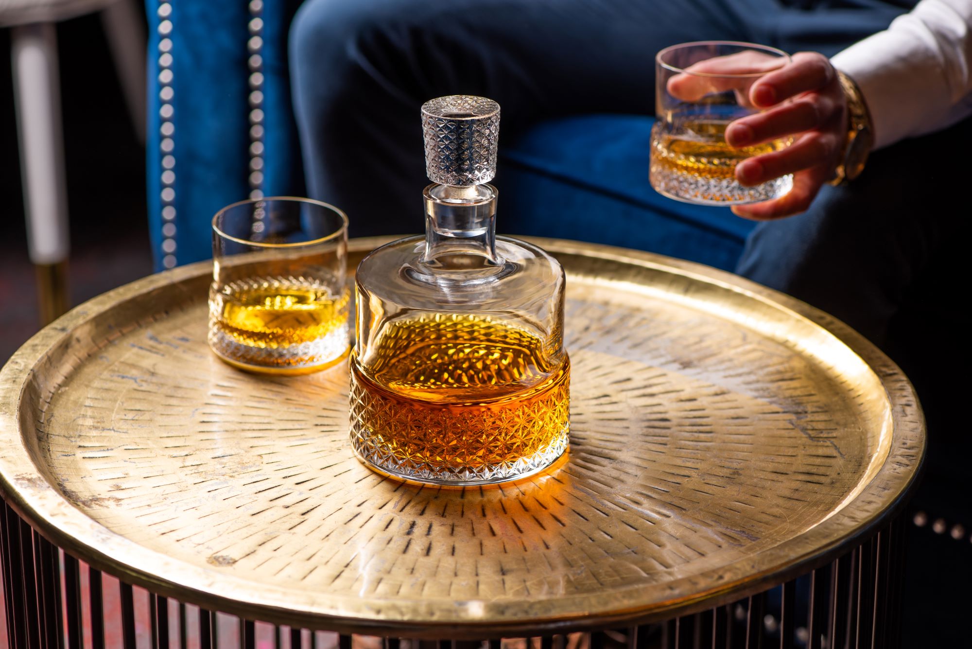 A Rona cut crystal whisky decanter and two matching tumblers containing amber spirit, arranged on a gold tray with a blue velvet armchair visible in the background