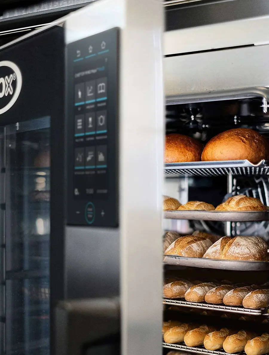 Multiple trays of freshly baked bread loaves inside an open Unox BAKERTOP MIND.Maps oven, showing the touch control panel and loaded baking chamber