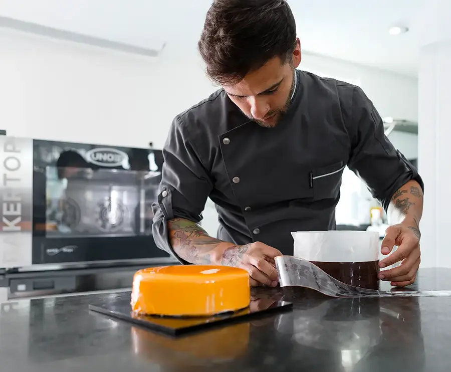 A professional pastry chef applying a chocolate acetate strip to a mirror-glazed entremet cake, with a Unox BAKERTOP oven visible in the background