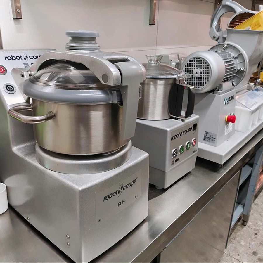 Robot Coupe R8 and R5 cutter mixers and a vegetable preparation machine installed on a stainless steel prep bench in a retail kitchen